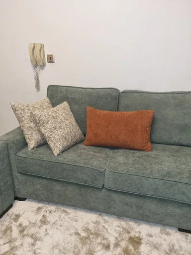 كنبة خضراء مع وسائد بلون الصدأ المميزة بجانب نافذة في منزل عميل بجدة